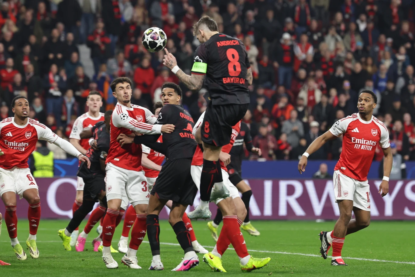 Piłkarz Bayernu Monachium w czarnym stroju z numerem 8 wyskakuje do piłki podczas meczu przeciwko zawodnikom Arsenalu ubranym w czerwono-białe stroje, otoczony przez kilku przeciwników przed bramką, w tle widać widownię stadionu.