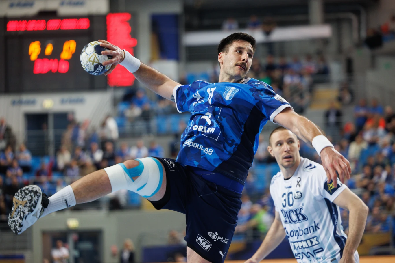 Abel Serdio Zawodnik w niebieskiej koszulce wykonuje dynamiczny wyskok z piłką podczas meczu piłki ręcznej, obok znajduje się obrońca drużyny przeciwnej w białym stroju, a w tle widoczna jest publiczność oraz elektroniczna tablica wyników.