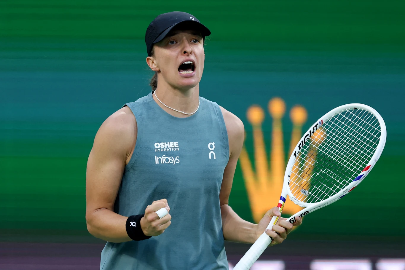 Tenisistka w sportowym stroju i czapce z daszkiem trzyma rakietę w jednej ręce i zaciska pięść w geście triumfu, wyrażając silne emocje podczas meczu na tle rozmazanego zielonego ekranu z logo zegarka Rolex.