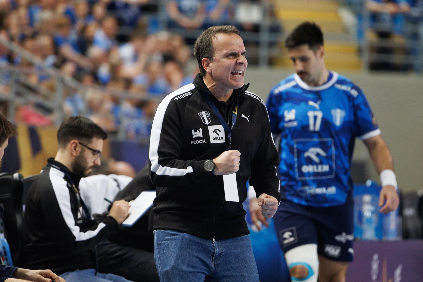 Xavier Sabate Trener w czarnej bluzie z logo Puma energicznie wydaje polecenia podczas meczu piłki ręcznej, obok niego zawodnik w niebieskim stroju z numerem 17 i opatrunkiem na kolanie, w tle rozmyta publiczność na trybunach.