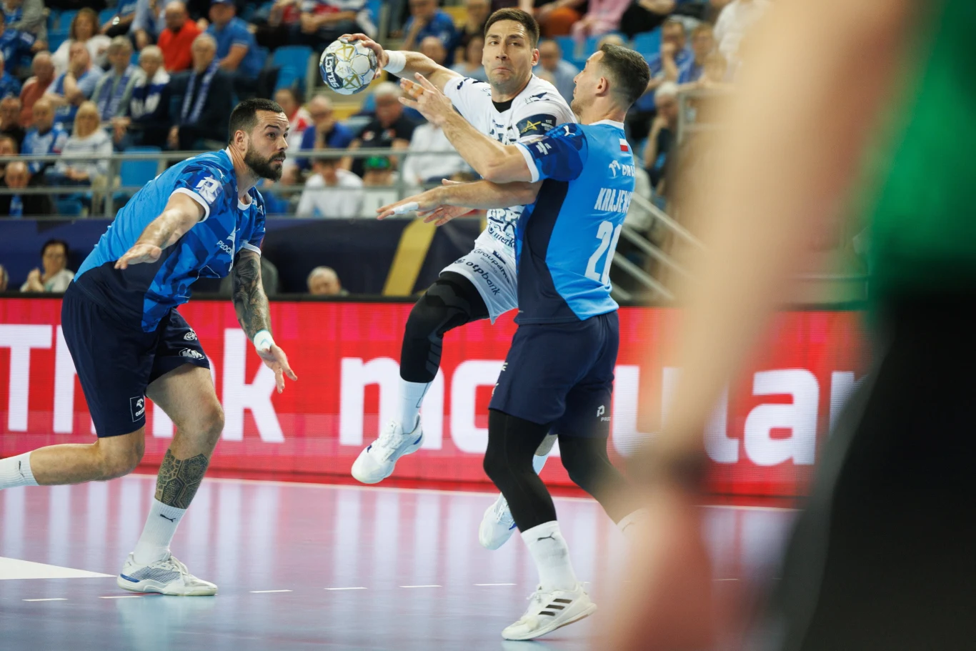 W akcji Lazar Kukić. Serb był najczęściej faulowanym zawodnikiem Picku Zawodnik w białym stroju wykonuje rzut w powietrzu podczas meczu piłki ręcznej, otoczony przez dwóch obrońców w niebieskich koszulkach, widownia w tle uważnie obserwuje akcję.