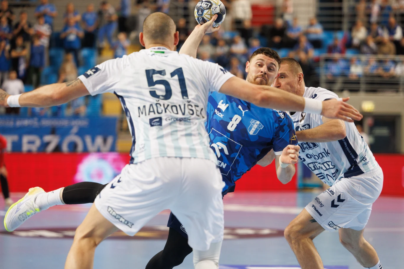 Mitja Janc Dynamiczna akcja podczas meczu piłki ręcznej, zawodnik w niebieskiej koszulce próbuje rzucić piłkę na bramkę, walcząc z dwoma obrońcami w białych strojach, z wyraźną ekspresją rywalizacji i napięcia sportowego.