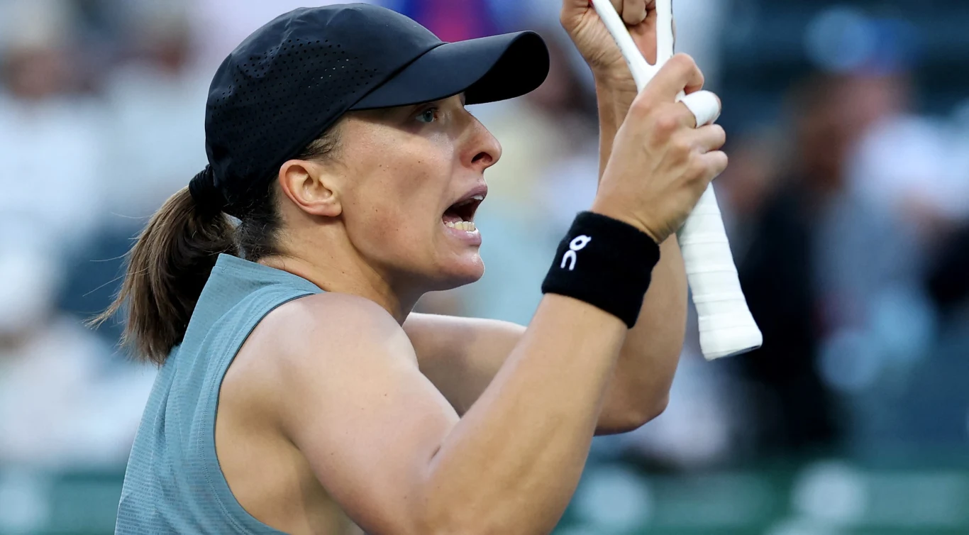 Kobieta ubrana w sportową odzież i czapkę z daszkiem trzyma zaciśniętą dłoń na rakiecie tenisowej, wyrażając silne emocje podczas meczu.