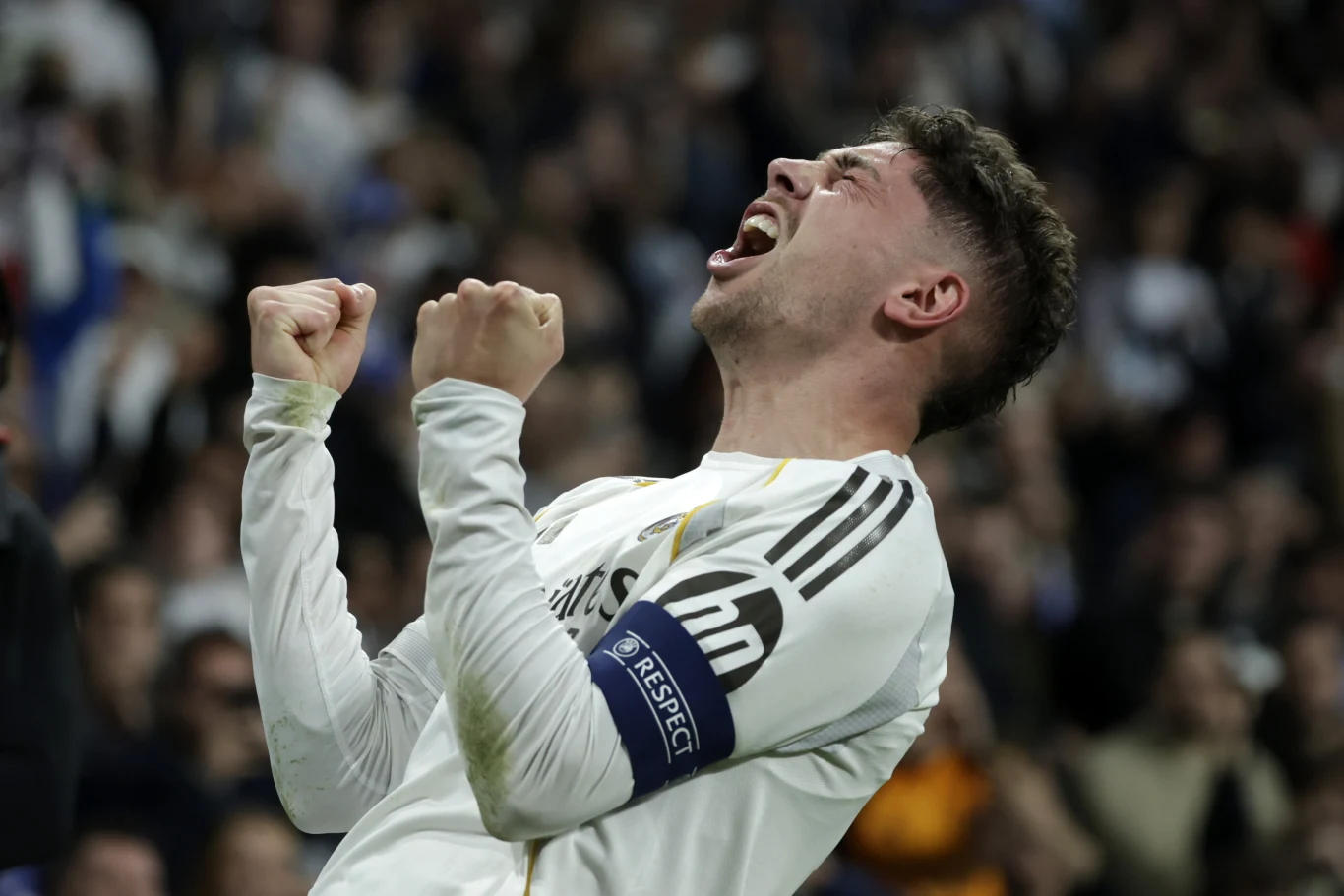 Piłkarz w białej koszulce drużyny Real Madryt z opaską kapitańską na ramieniu, zaciśniętymi pięściami i wyrażający silne emocje radości, otoczony rozmazanym tłumem na stadionie.