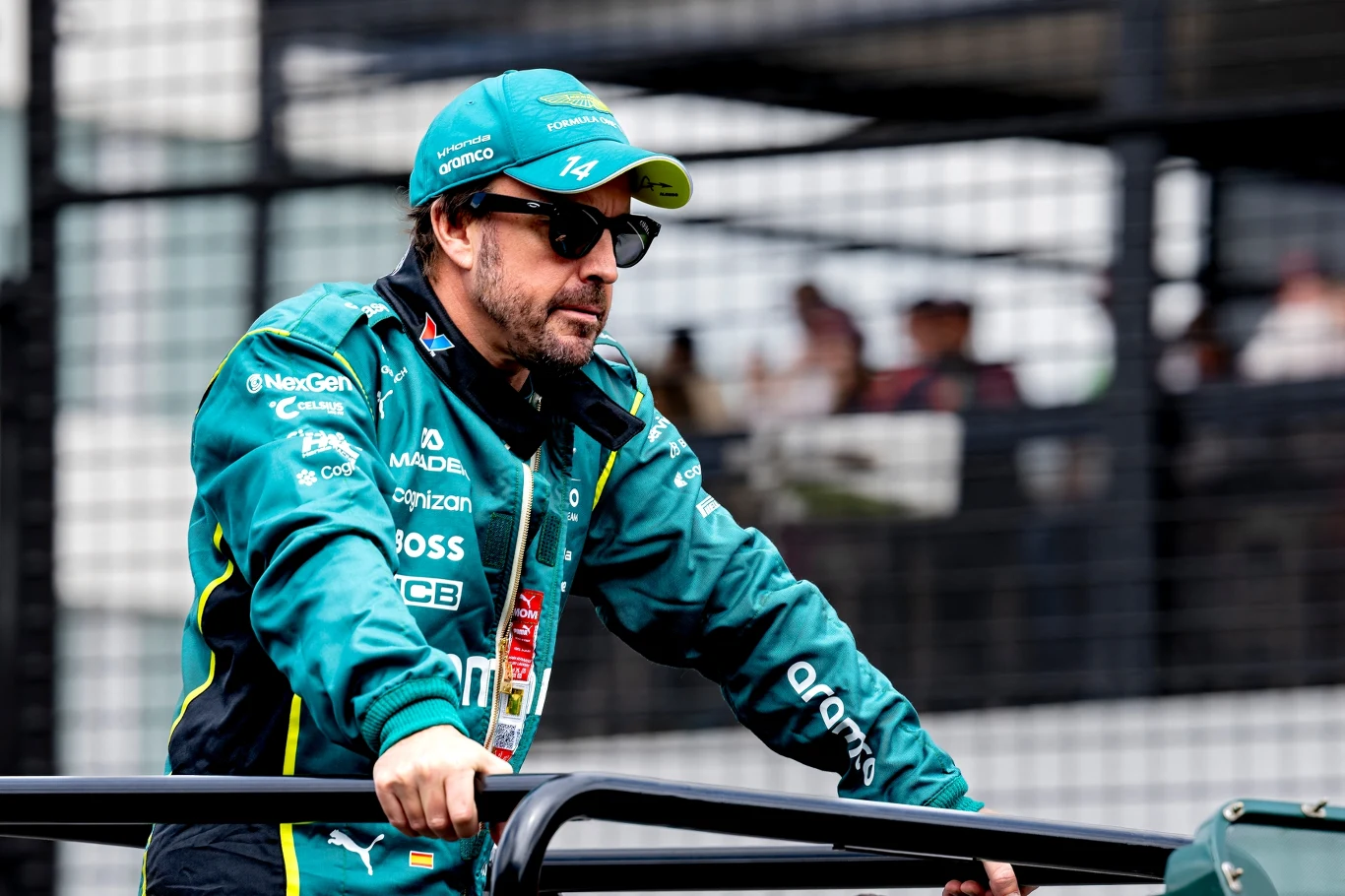 Mężczyzna w zielonej kurtce zespołu wyścigowego Aston Martin i czapce z daszkiem prowadzi pojazd, ma na sobie okulary przeciwsłoneczne, w tle widoczna metalowa siatka ogrodzeniowa i rozmazane sylwetki ludzi.