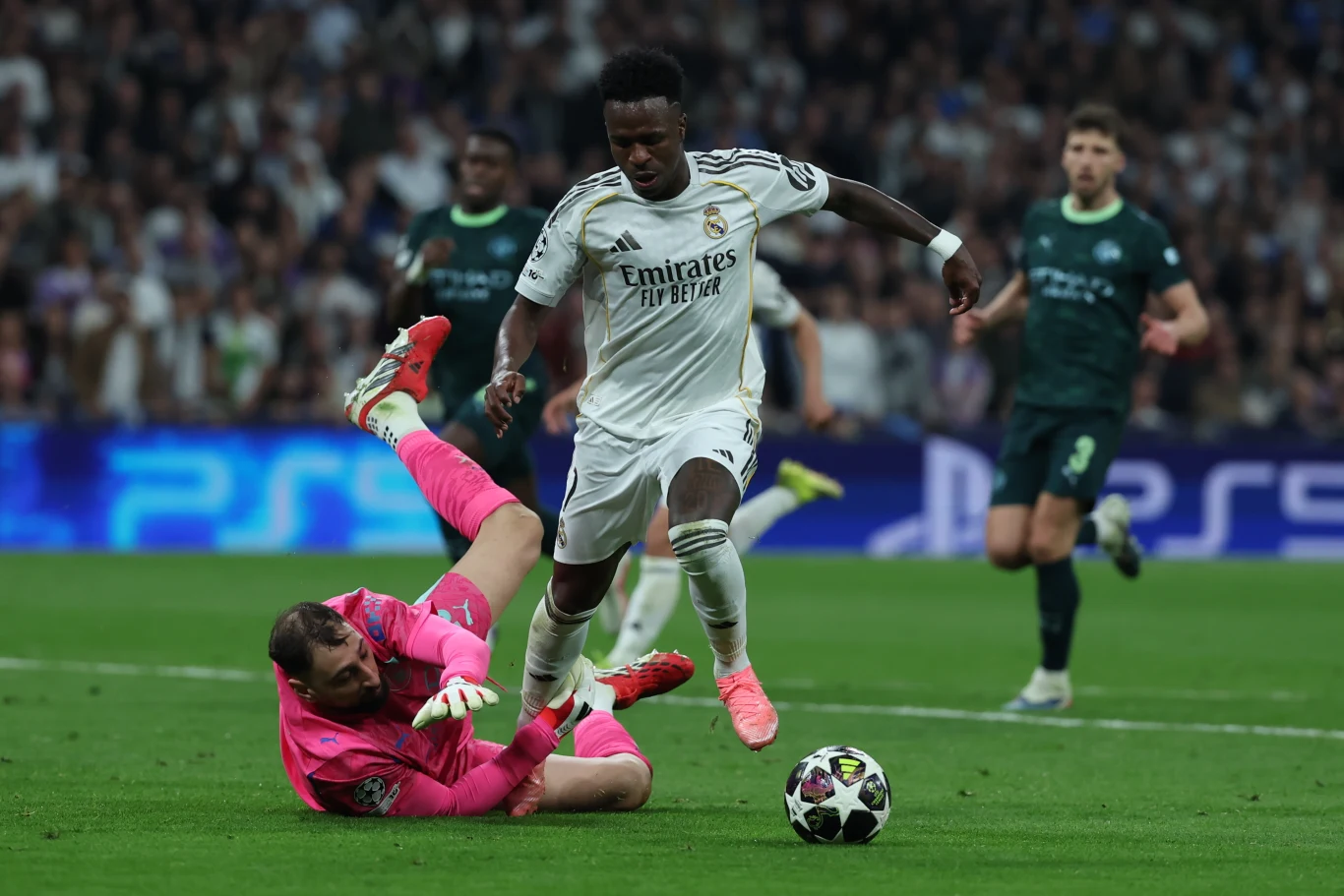 Piłkarz w białym stroju z piłką przy nodze próbuje minąć bramkarza leżącego na murawie w różowym komplecie podczas meczu piłki nożnej na stadionie pełnym kibiców.