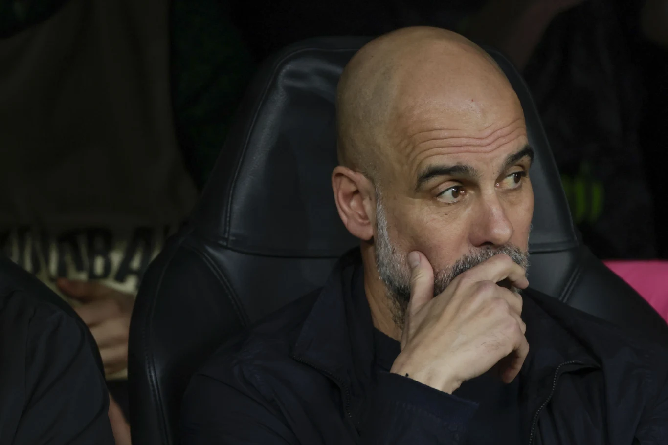 Łysy mężczyzna z brodą w ciemnej kurtce siedzi na czarnym fotelu, opiera brodę na dłoni i wygląda zamyślony.