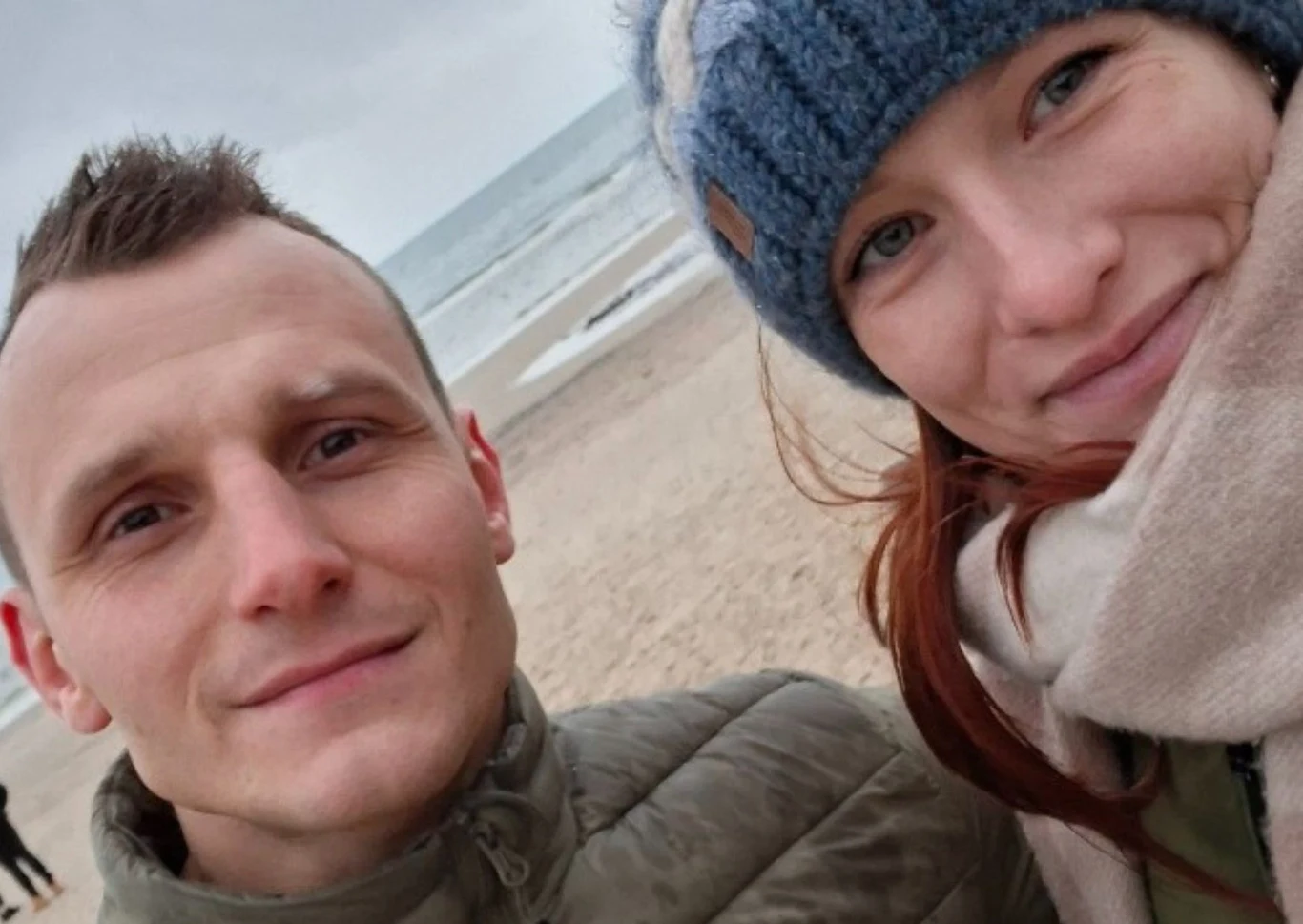 Para uśmiechniętych osób ubranych w ciepłe kurtki i szalik, stojąca blisko siebie na piaszczystej plaży z widocznym w tle morzem i pochmurnym niebem.