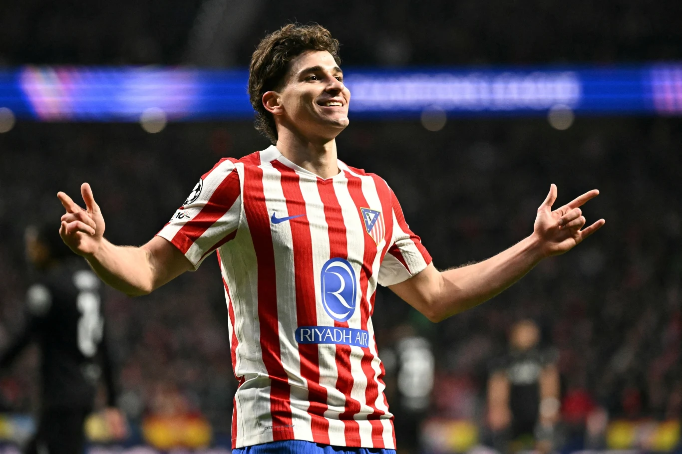 Julian Alvarez Piłkarz w czerwono-białej koszulce Atletico Madryt celebruje zdobytego gola, rozkładając ręce i uśmiechając się szeroko na tle rozmytego widowni stadionowej.