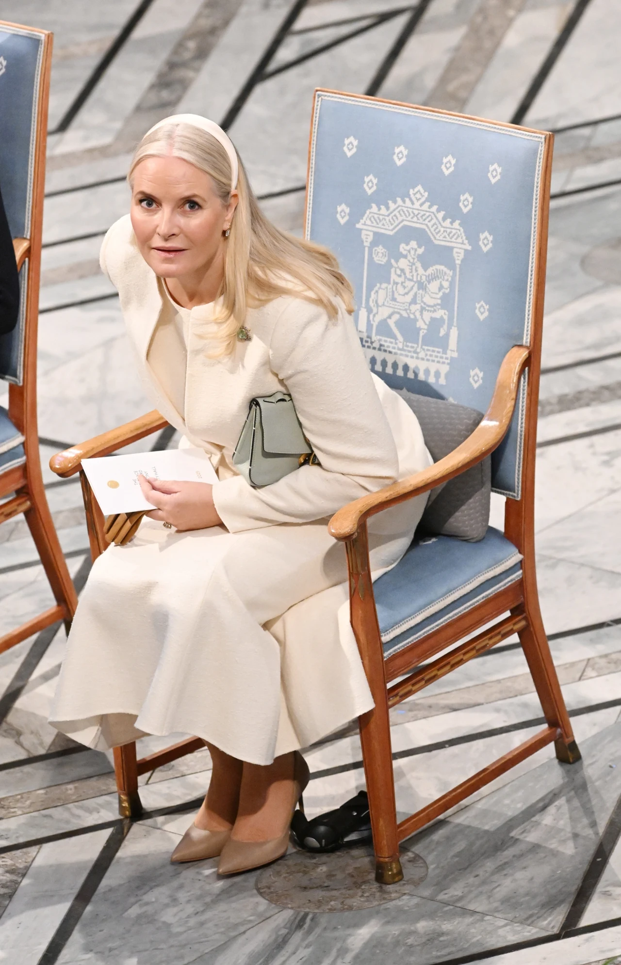 Kobieta w jasnym eleganckim stroju siedzi na ozdobionym niebieskim krześle z drewnianymi podłokietnikami, trzyma kopertę i torebkę, spoglądając w górę; krzesło dekorowane białym wzorem przedstawiającym postać na koniu.