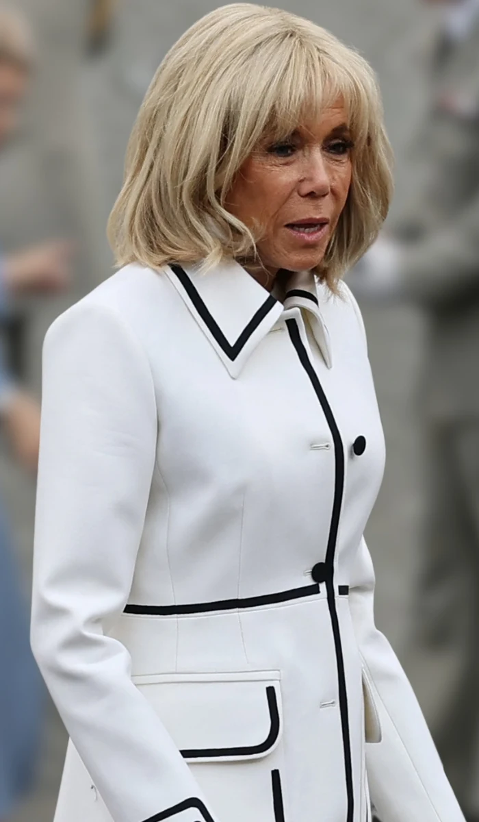 Kobieta o blond włosach ubrana w elegancki biały płaszcz z czarnymi lamówkami, stojąca na tle rozmazanych sylwetek innych osób ubraných w szarości.