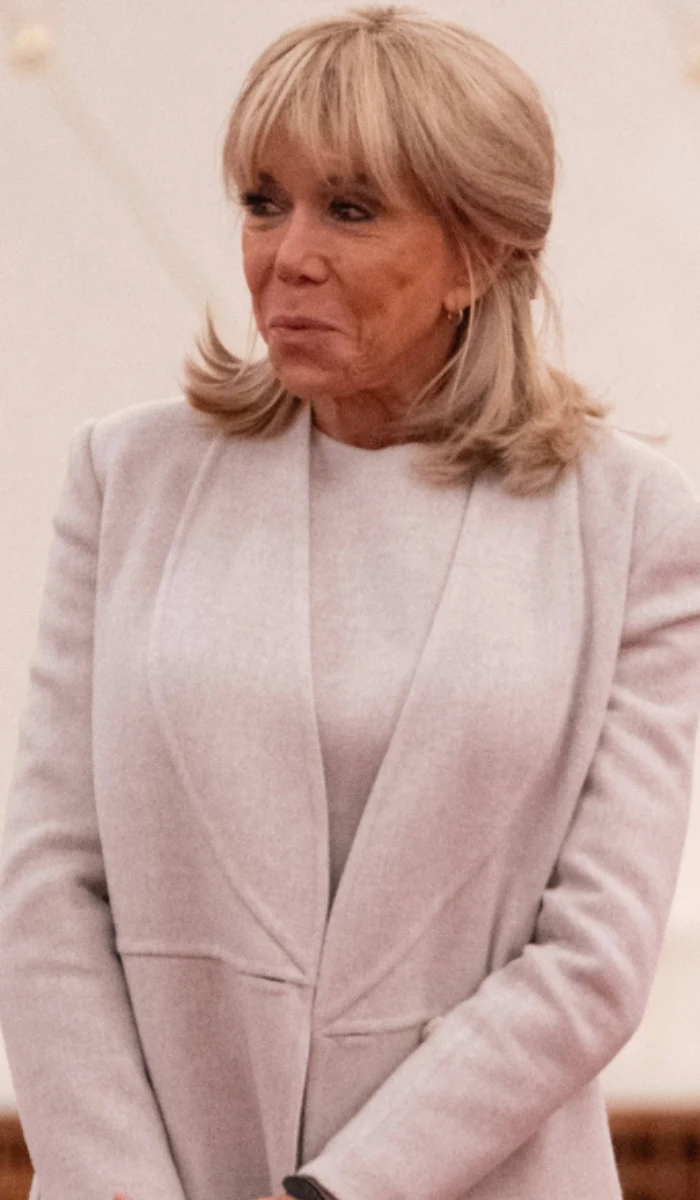 Kobieta o jasnych włosach w półdługiej fryzurze ubrana w elegancki, jasny żakiet i sweter, z delikatnym makijażem oraz złotymi kolczykami, patrząca lekko w bok w spokojnym, zamkniętym otoczeniu.