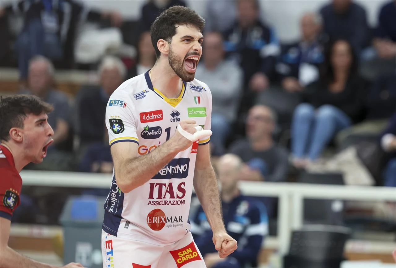 Siatkarz w białym stroju z logo ITAS świętuje zdobycie punktu, zaciśnięta pięść i wyrazista radość na twarzy podkreślają emocje sportowej rywalizacji, w tle widoczni kibice oraz inny zawodnik okazujący entuzjazm.