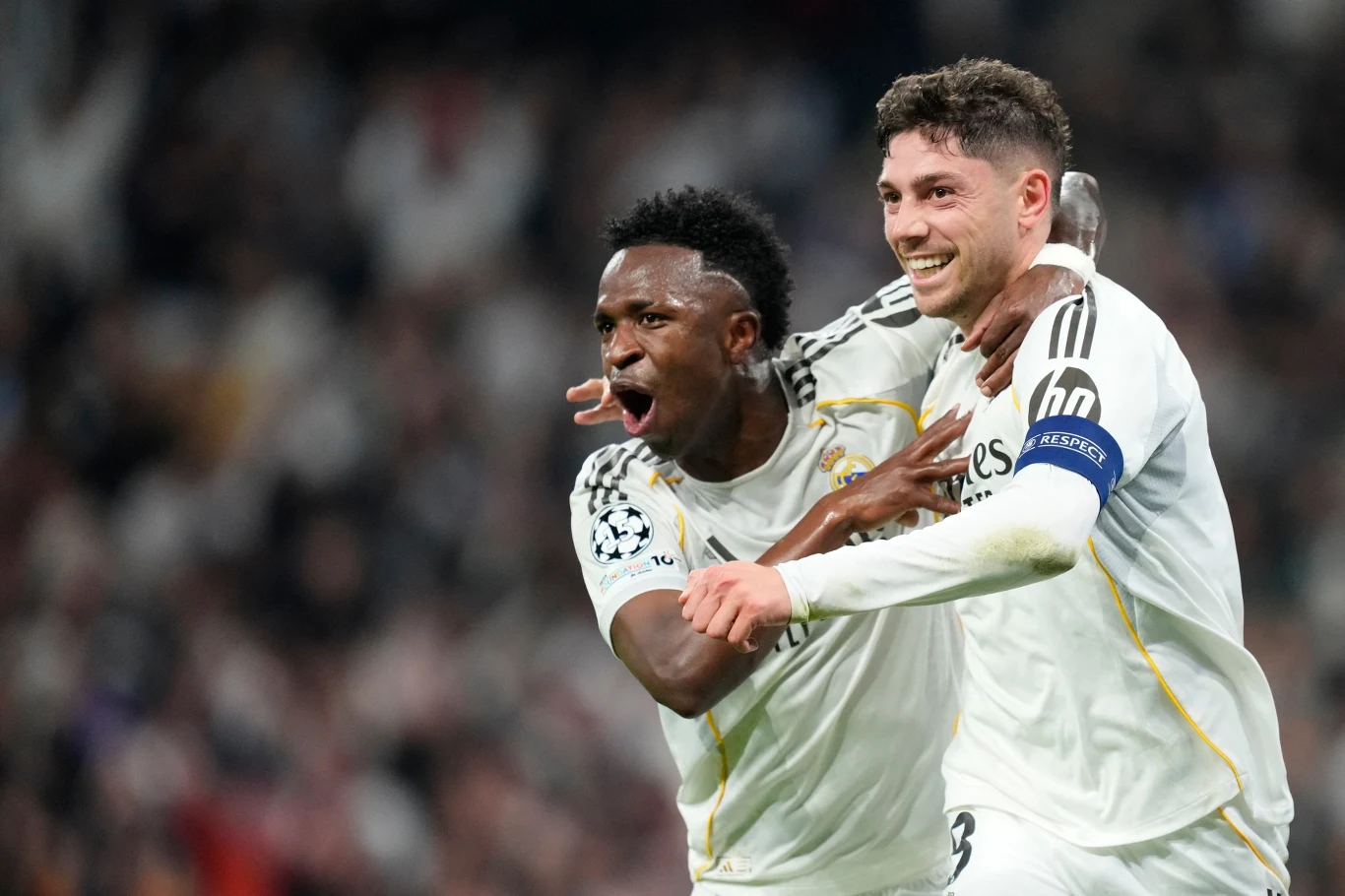 Dwóch piłkarzy drużyny Realu Madryt w białych strojach świętuje zdobycie gola na boisku, jeden z nich wyraża ogromną radość, a drugi obejmuje kolegę z zespołu z uśmiechem na twarzy.