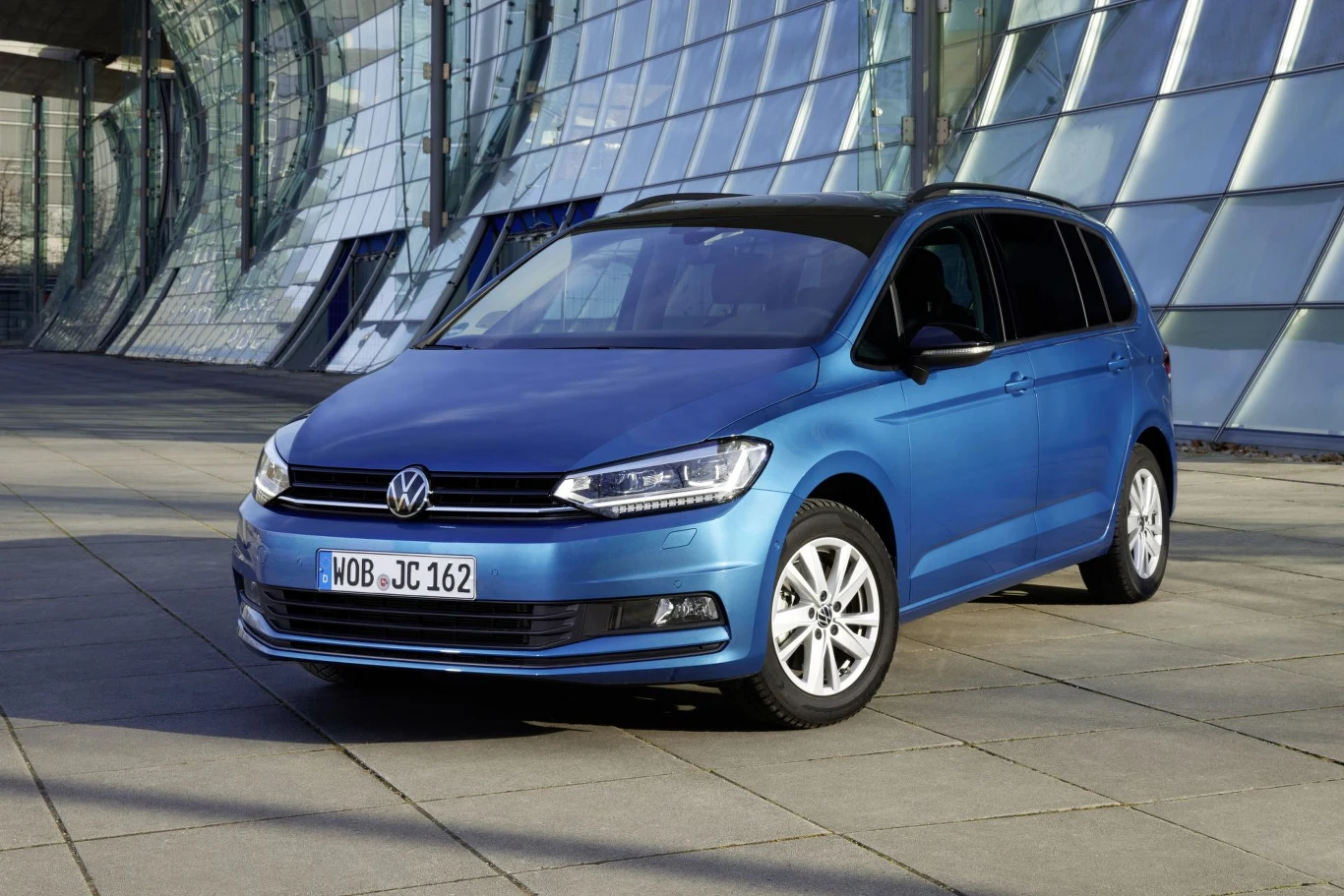 niebieski samochód Volkswagen zaparkowany na tle nowoczesnego, przeszklonego budynku o zakrzywionej fasadzie