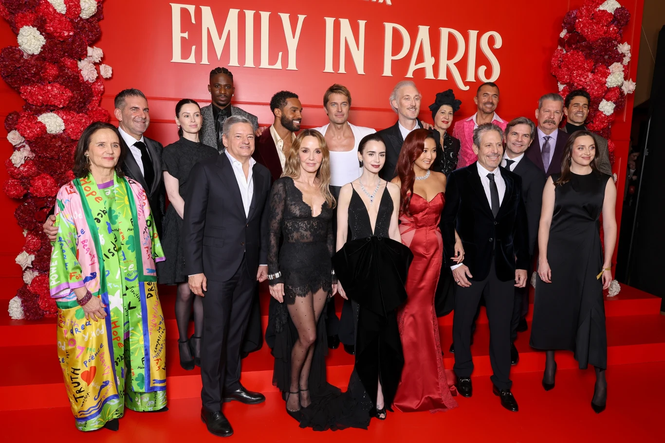 Grupa elegancko ubranych osób pozuje na tle czerwonej ścianki ozdobionej kwiatami, z dużym napisem 'Emily in Paris'.