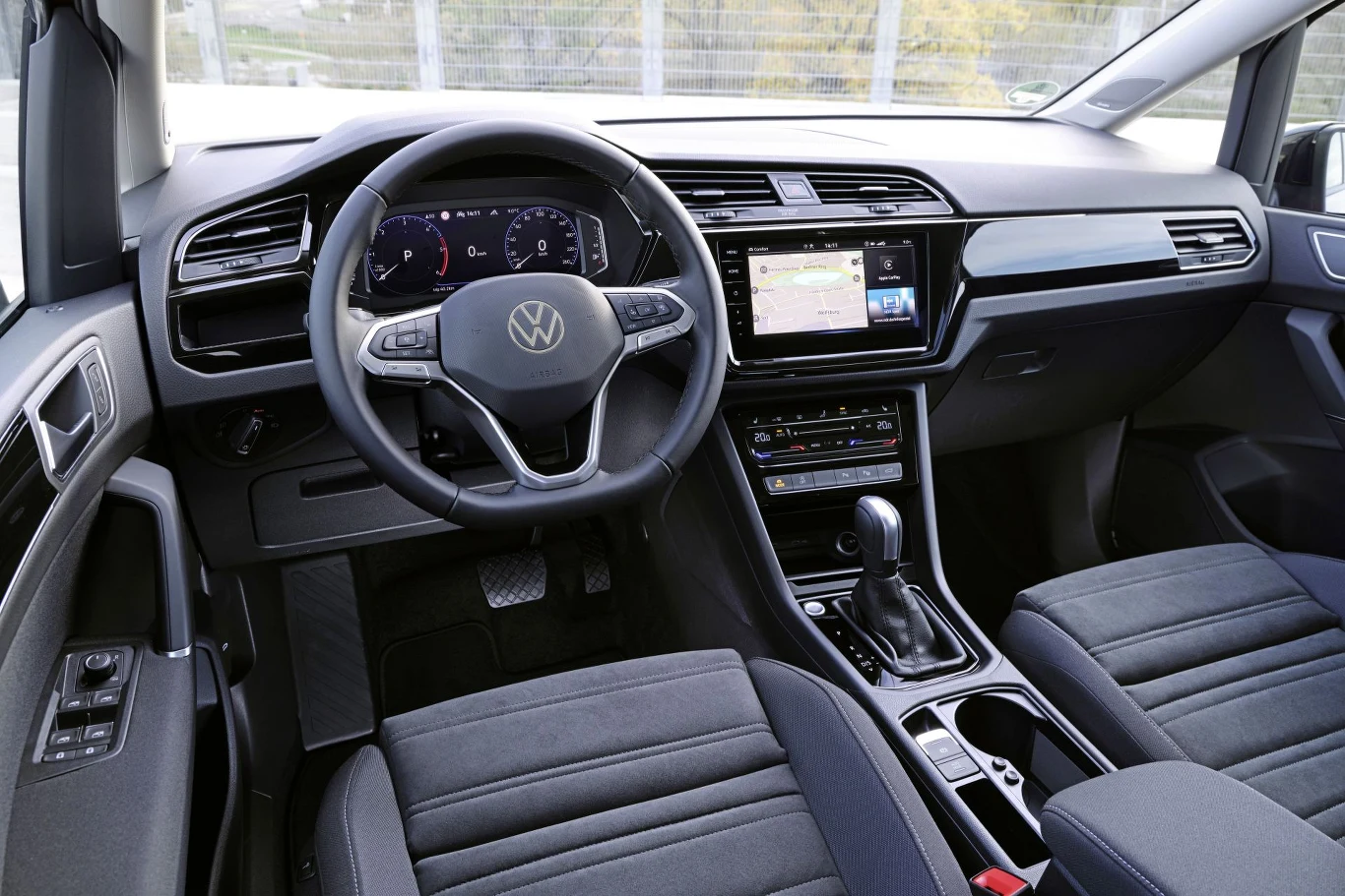 Wnętrze samochodu marki Volkswagen z cyfrowym panelem zegarów, dużym ekranem systemu multimedialnego pośrodku deski rozdzielczej, automatyczną skrzynią biegów oraz fotelami pokrytymi ciemną tapicerką materiałową.