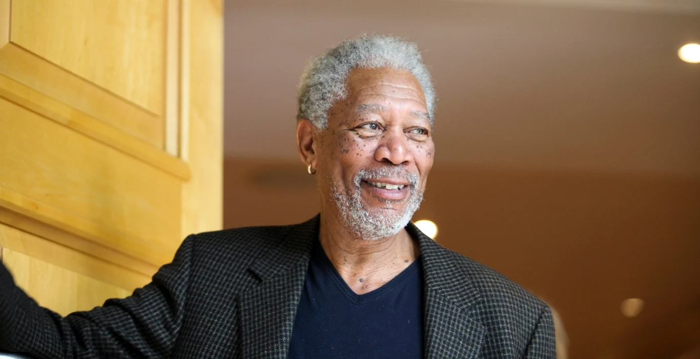 Morgan Freeman Starszy mężczyzna z siwymi włosami i brodą, ubrany w ciemny T-shirt oraz marynarkę w drobną kratkę, stoi przy jasnych drewnianych drzwiach i uśmiecha się delikatnie, patrząc w bok.