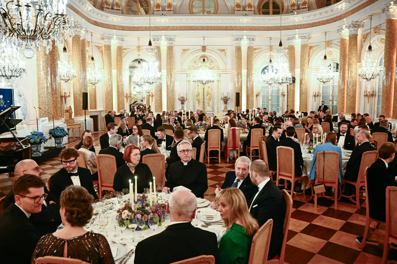 Uroczysta kolacja w eleganckiej sali balowej z dużą liczbą gości ubranych w wieczorowe stroje siedzących przy okrągłych stołach ozdobionych świecami i kwiatami; wysokie zdobione sufity, liczne żyrandole i bogata architektura wnętrza podkreślają podnios...