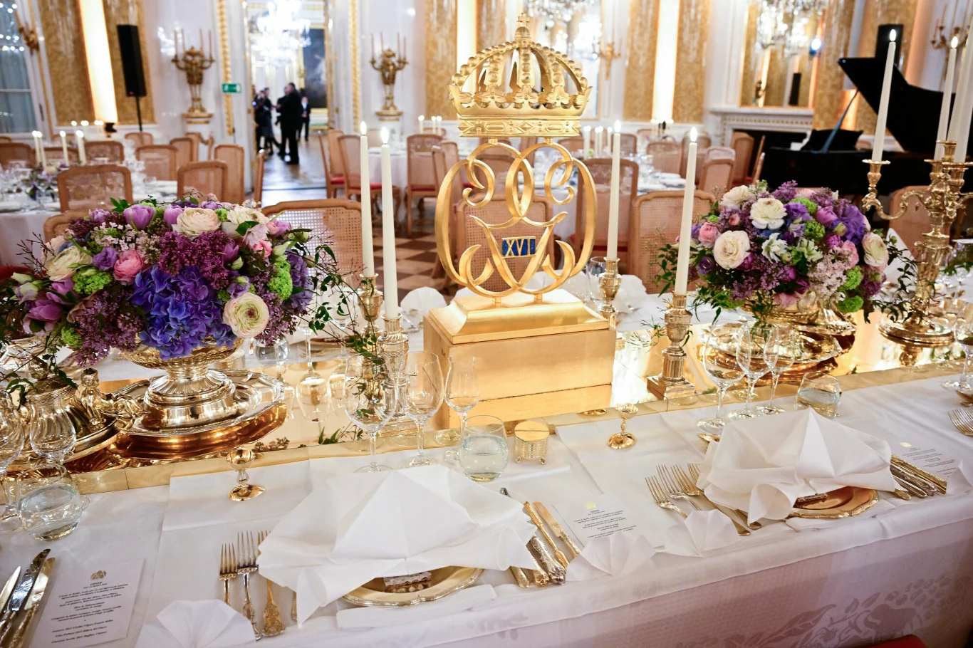 Bogato zdobiony stół bankietowy ustawiony w eleganckiej, złoconej sali pałacowej, udekorowany dużą pozłacaną koroną, świecznikami, bukietami różnokolorowych kwiatów oraz starannie złożonymi serwetkami i wykwintną zastawą.