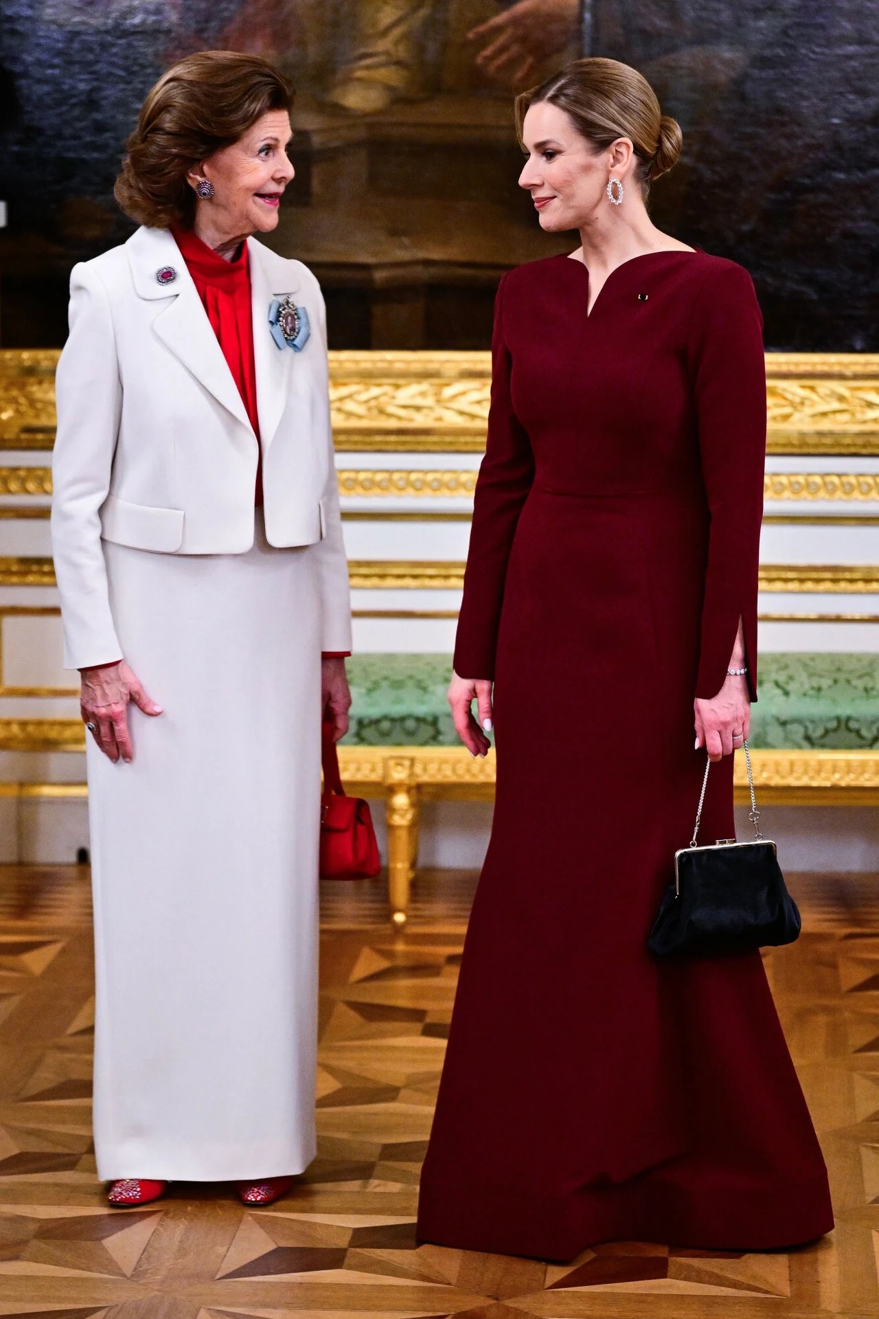 Dwie elegancko ubrane kobiety stoją na parkiecie w bogato zdobionym wnętrzu; jedna jest ubrana w długą, bordową suknię i trzyma czarną torebkę, druga nosi białą garsonkę z czerwonym golfem oraz broszką na marynarce, obie patrzą na siebie z uśmiechem.