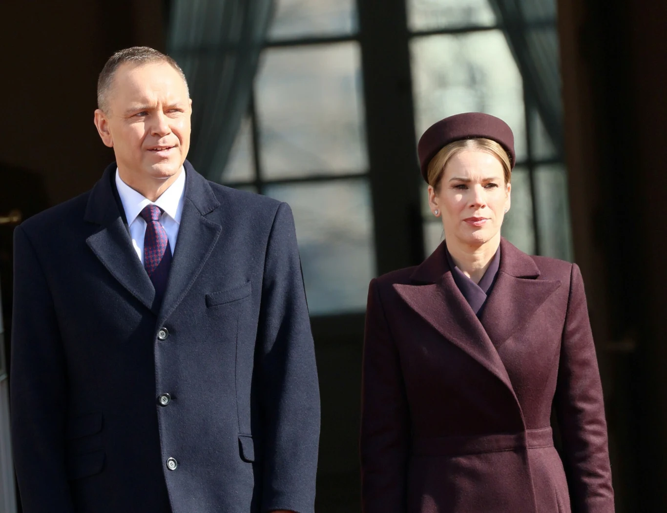 Dwie elegancko ubrane osoby stoją obok siebie, mężczyzna w ciemnym płaszczu i krawacie, kobieta w bordowym płaszczu oraz dopasowanym kapeluszu, na tle dużego okna z zasłonami.