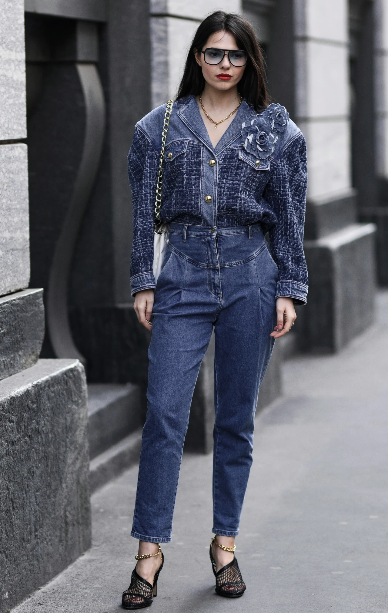 Kobieta ubrana w stylizację typu denim-on-denim: granatowa kurtka jeansowa z ozdobną aplikacją na ramieniu oraz wysokie, dopasowane spodnie jeansowe, uzupełnione czarnymi sandałami na obcasie, małą torebką na łańcuszku oraz dużymi okularami przeciwsłon...