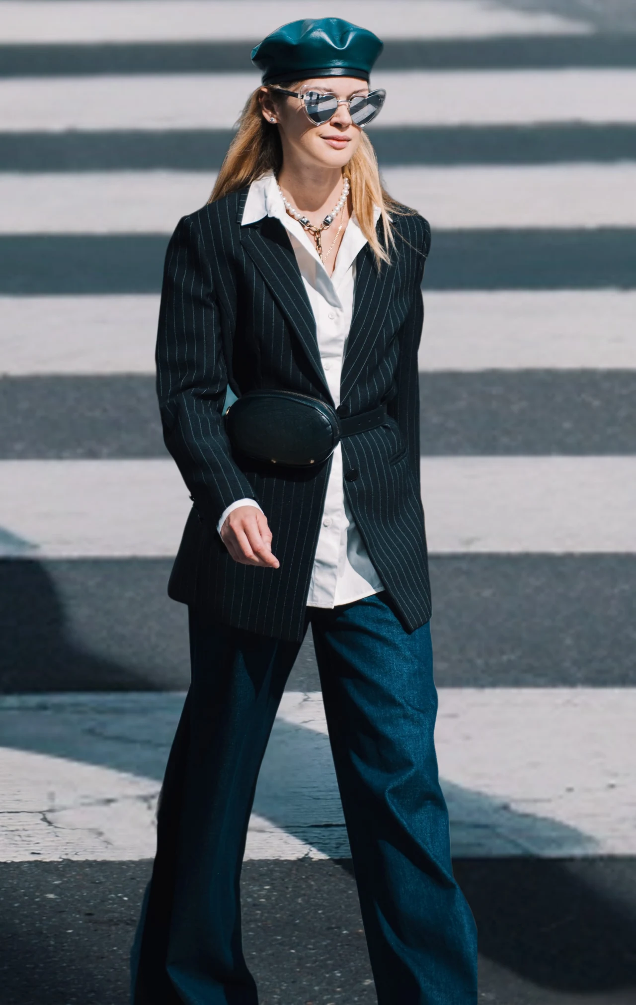 Kobieta w stylowej, casualowej stylizacji idzie przez pasy; ma na sobie granatowy beret, ciemne okulary przeciwsłoneczne, czarną marynarkę w prążki, białą koszulę, szerokie jeansy oraz saszetkę przewieszoną przez talię.