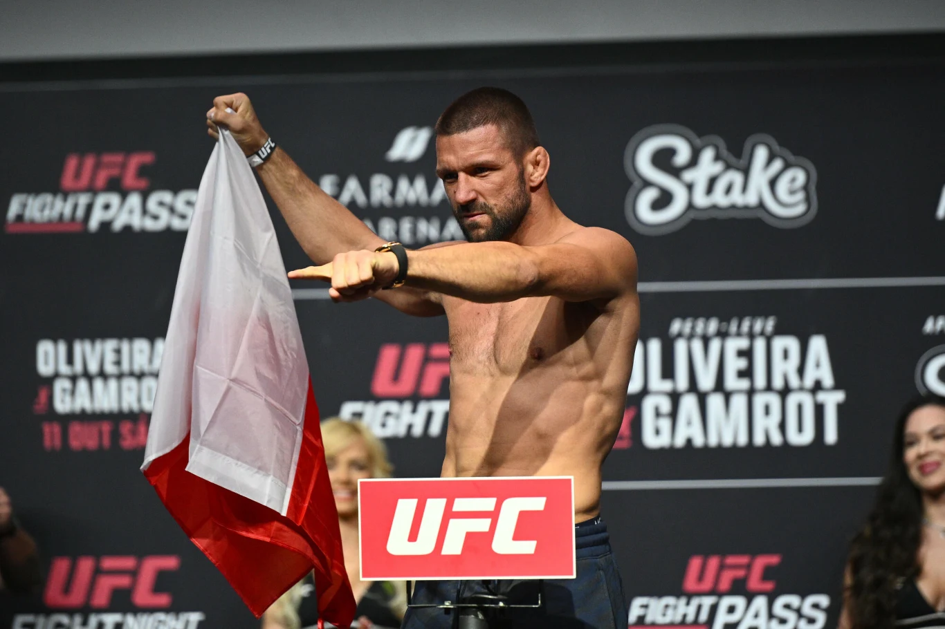Zawodnik MMA na tle tablicy z logo UFC stoi na wadze, trzyma w jednej ręce polską flagę i wskazuje przed siebie, prezentując umięśnioną sylwetkę.