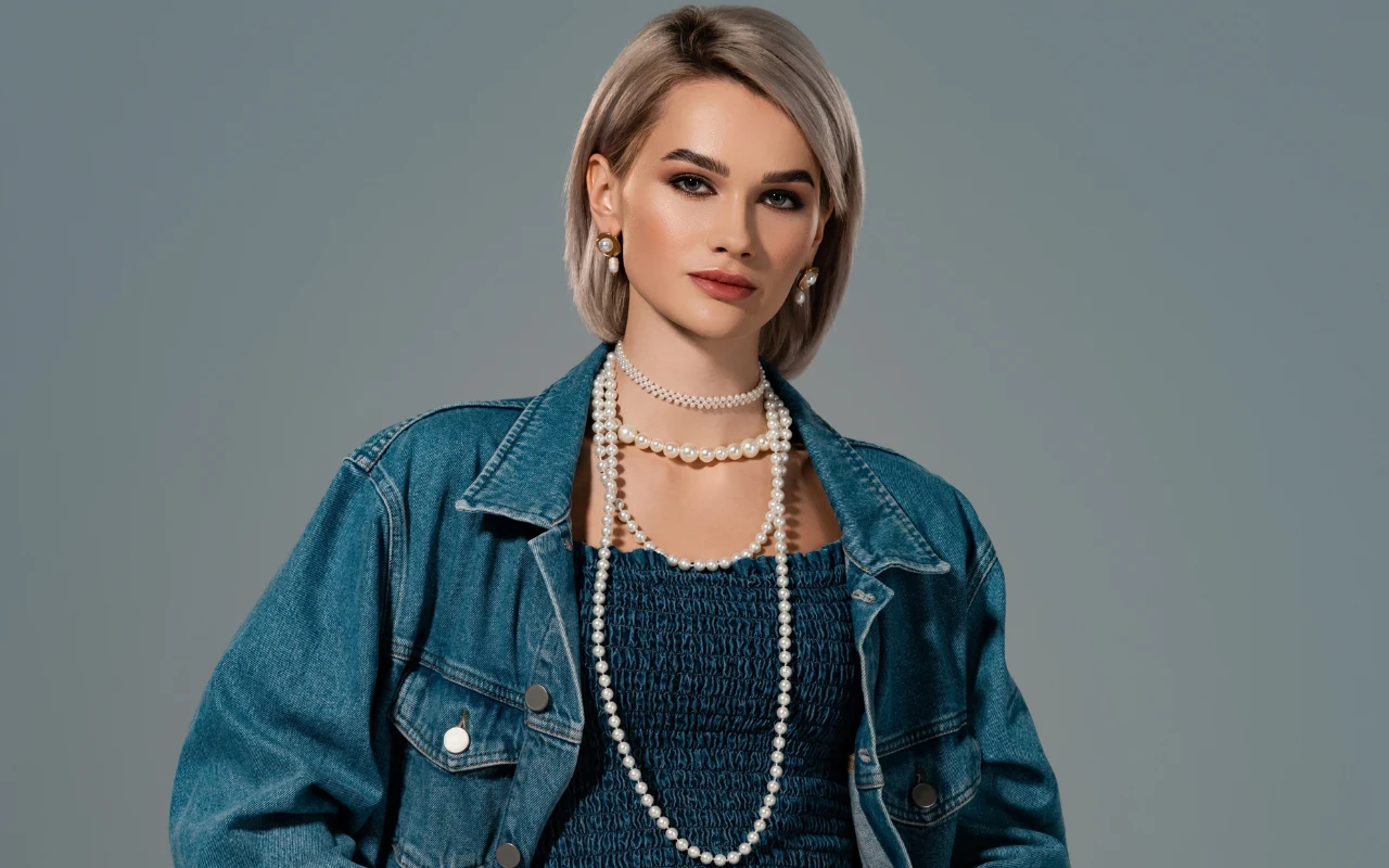 Kobieta z krótkimi blond włosami ubrana w ciemnoniebieską sukienkę i jeansową kurtkę, ozdobiona długim naszyjnikiem z pereł oraz perłowymi kolczykami, patrząca spokojnie w kierunku obiektywu na neutralnym tle.