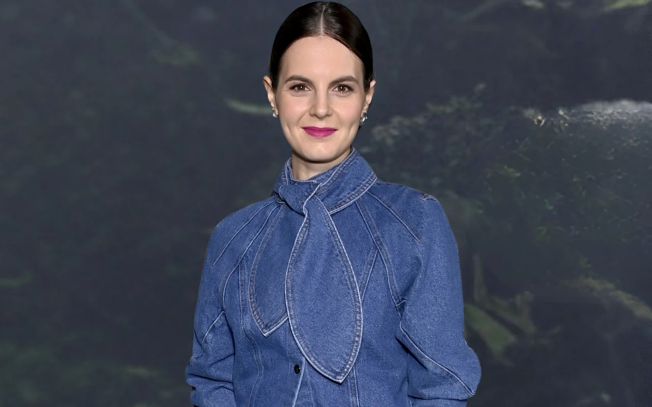 Kobieta z ciemnymi, gładko zaczesanymi włosami, ubrana w niebieską kurtkę dżinsową z ozdobnym wiązaniem pod szyją, stoi na tle ciemnej, lekko rozmytej scenerii przypominającej naturalne otoczenie.