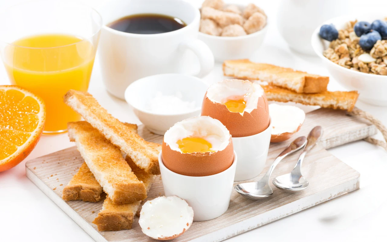 Jajka na miękko w kieliszkach, podane z tostami, kawa, sok pomarańczowy, świeża połówka pomarańczy, miseczka cukru oraz jogurt z musli i borówkami, wszystko na jasnym tle śniadaniowym.