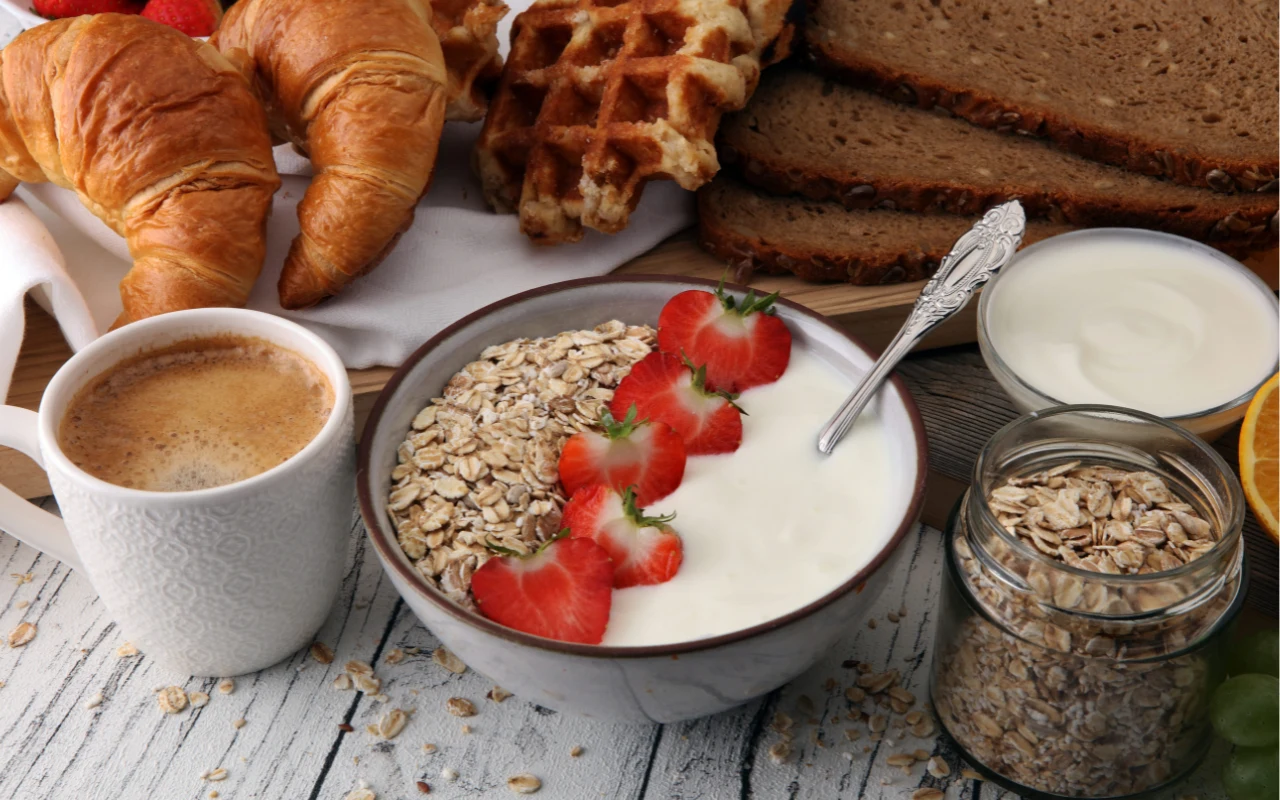 Miska jogurtu z płatkami owsianymi i plasterkami truskawek, obok kubek kawy, croissanty, gofry, kromki chleba żytniego oraz dodatkowe płatki owsiane i jogurt w małych naczyniach.