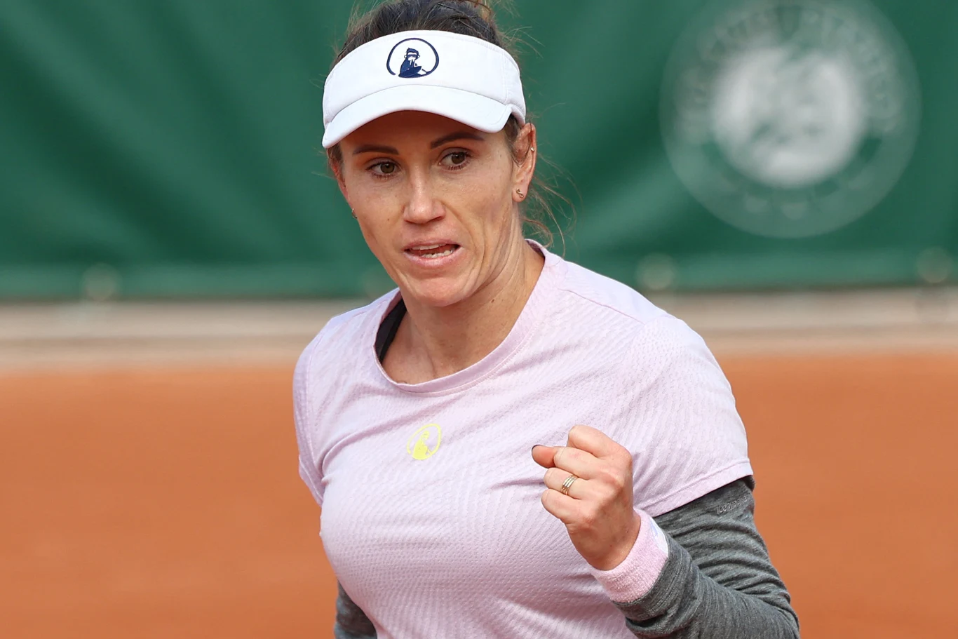 Tenisistka w jasnej koszulce i białej czapce z daszkiem ściska zaciśniętą pięść w geście triumfu, stojąc na korcie ziemnym.