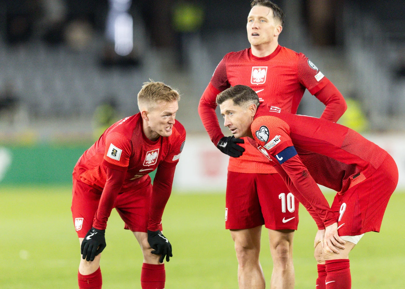 Trzech piłkarzy reprezentacji Polski w czerwonych strojach omawia strategię podczas meczu, wykazując skupienie i zaangażowanie.