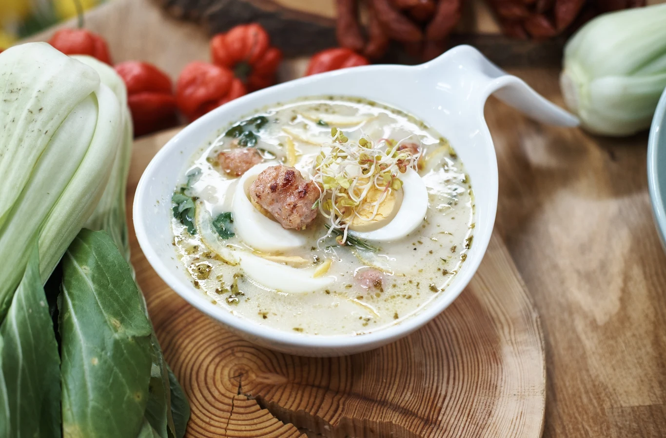 Biała zupa z kawałkami kiełbasy, jajkiem na twardo oraz kiełkami, podana w eleganckiej misce, obok świeża kapusta pak choi i czerwone papryczki w tle.