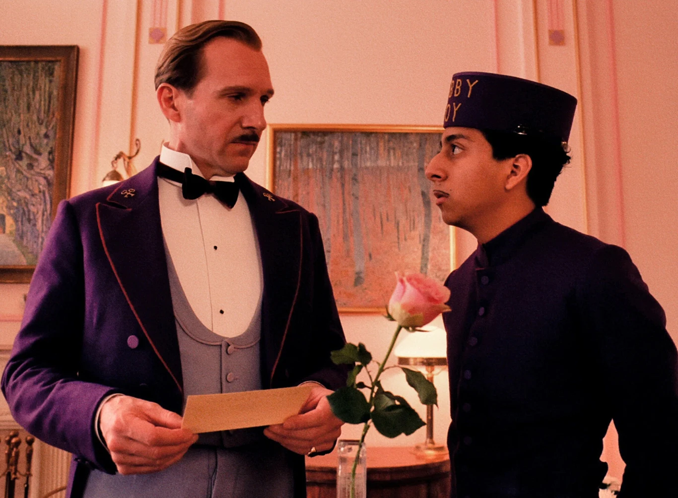 "Grand Budapest Hotel" Dwóch mężczyzn w eleganckich fioletowych uniformach hotelowych stoi w ozdobnym, jasnoróżowym pomieszczeniu; jeden z nich trzyma kopertę, a drugi różę, oboje patrzą na siebie w skupieniu.