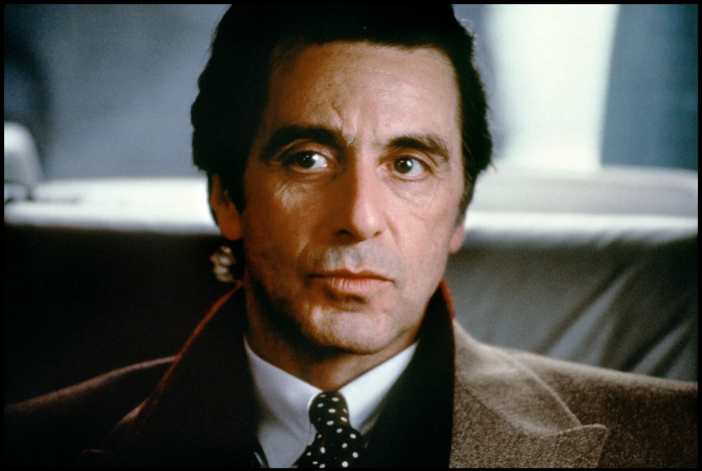Al Pacino w filmie "Zapach kobiety" Mężczyzna o poważnym wyrazie twarzy siedzi w samochodzie, ubrany elegancko w brązowy płaszcz, białą koszulę i krawat w kropki.