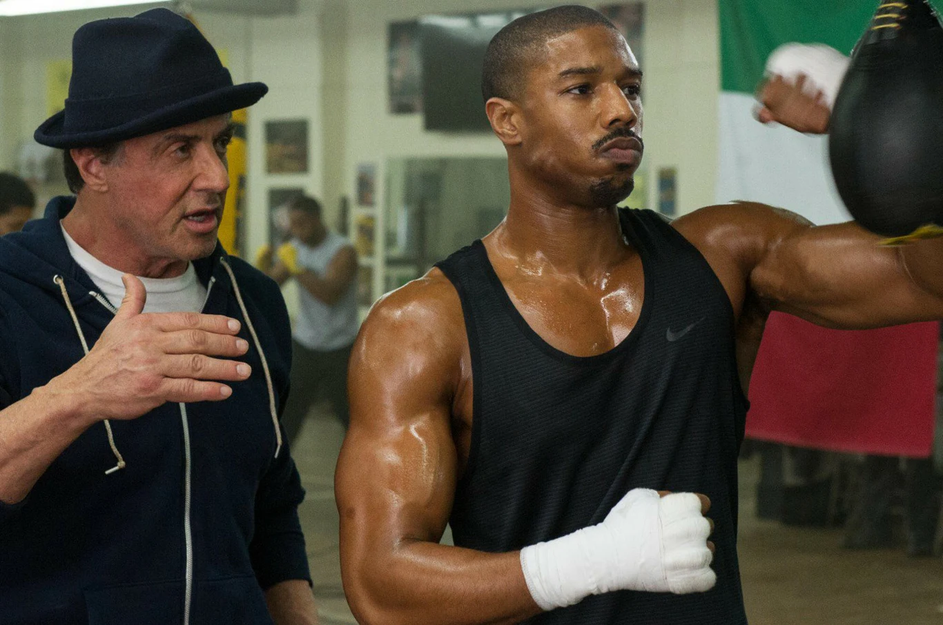 Sylvester Stallone i Michael B. Jordan w filmie "Credd. Narodziny legendy" Dwóch mężczyzn w siłowni bokserskiej, jeden w czarnym bezrękawniku ćwiczy uderzenia w worek treningowy, drugi w ciemnej czapce i rozpinanej bluzie gestykuluje i udziela wskazówek.