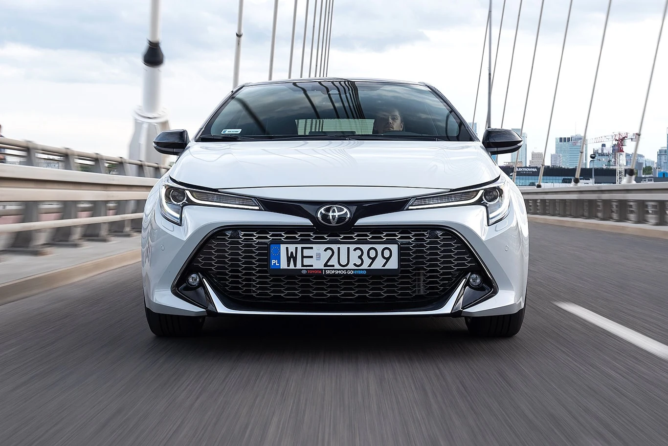 Biała Toyota Corolla z widocznym z przodu dużym, czarnym grillem i nowoczesnymi reflektorami, poruszająca się po moście z wyraźnymi stalowymi linami w tle