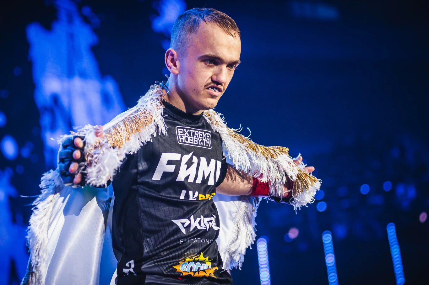 Mateusz "Mini Majk" Krzyżanowski Młody mężczyzna w sportowym stroju z nadrukiem FAME, okryty połyskującym płaszczem z frędzlami, stoi w dynamicznej pozie na arenie, koncentrując wzrok przed siebie, gotowy do walki.