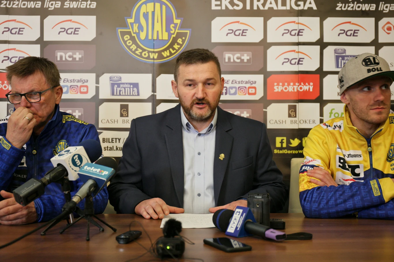 Trzech mężczyzn siedzących przy stole na konferencji prasowej klubu żużlowego Stal Gorzów Wielkopolski, pośrodku osoba oficjalna w garniturze, po bokach osoby w strojach klubowych, na stole mikrofony i sprzęt nagrywający, w tle ściana z logotypami spon...