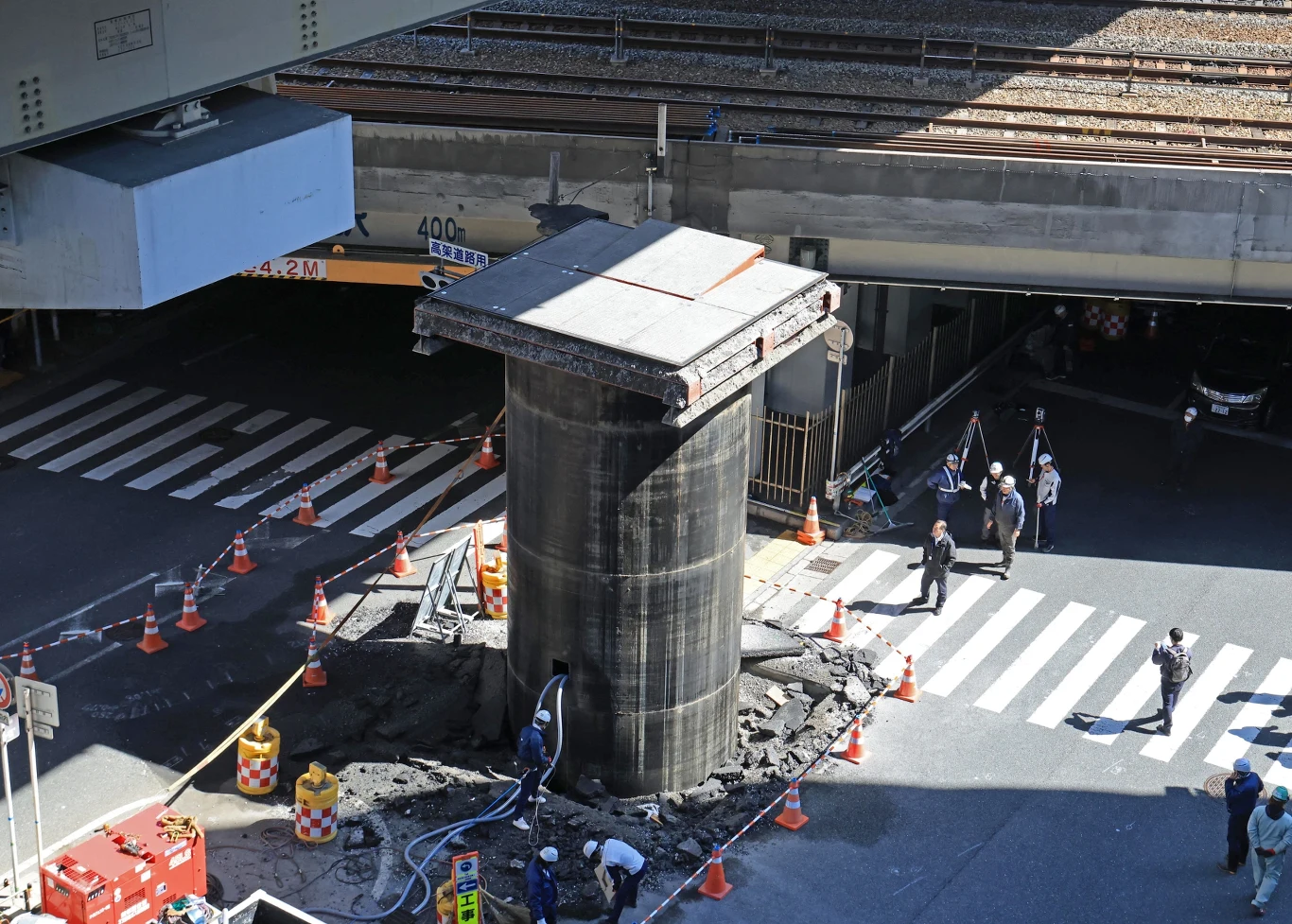 Duży betonowy filar otoczony pachołkami bezpieczeństwa i sprzętem budowlanym, robotnicy prowadzący prace konstrukcyjne na wyłączonym z ruchu skrzyżowaniu, w tle przejazd kolejowy nad ulicą oraz piesi obserwujący sytuację z bezpiecznej odległości.