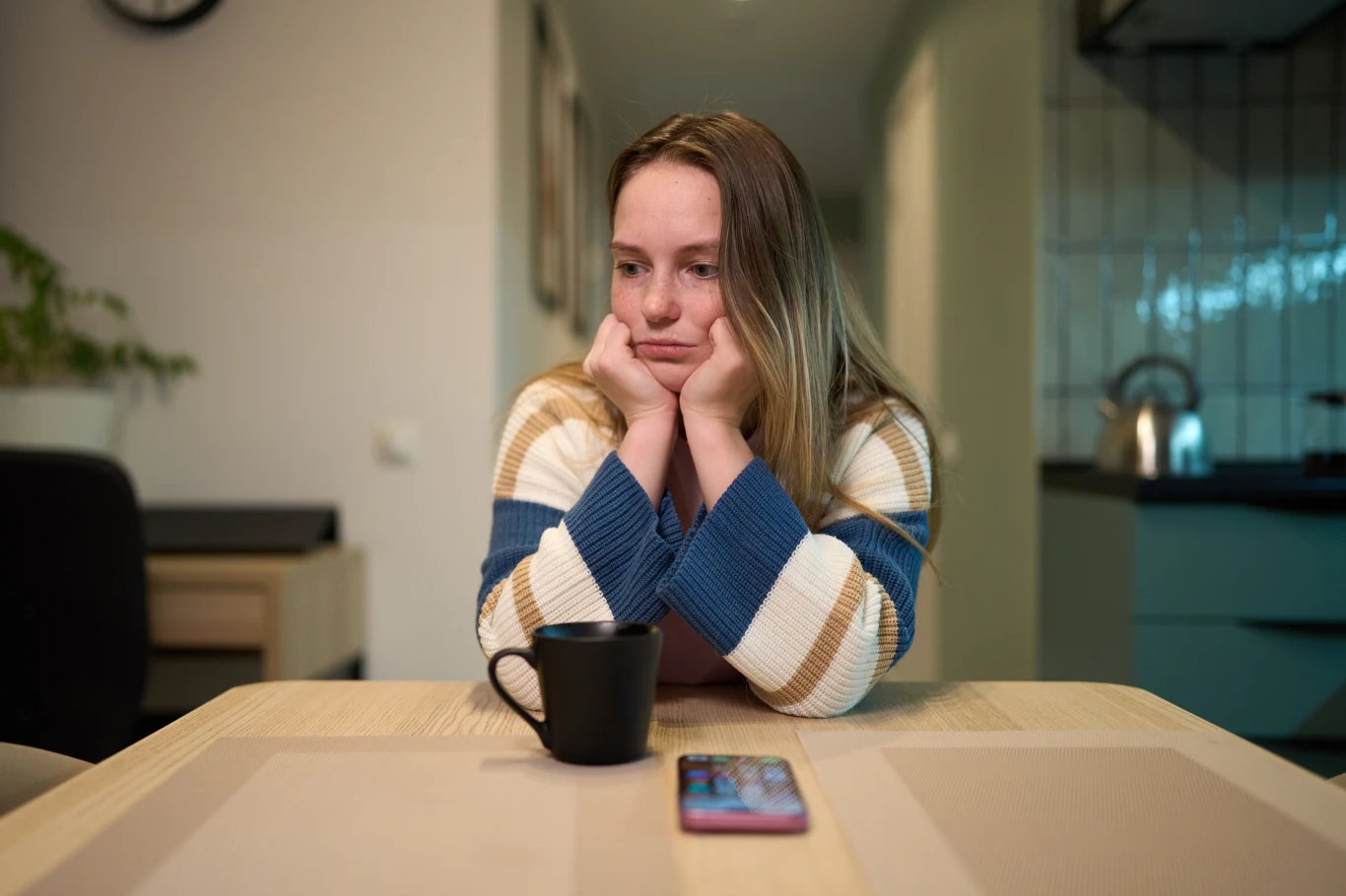 Kobieta siedzi przy stole z głową opartą na dłoniach, wyraźnie zamyślona lub zmartwiona; przed nią stoi czarny kubek oraz leży smartfon.