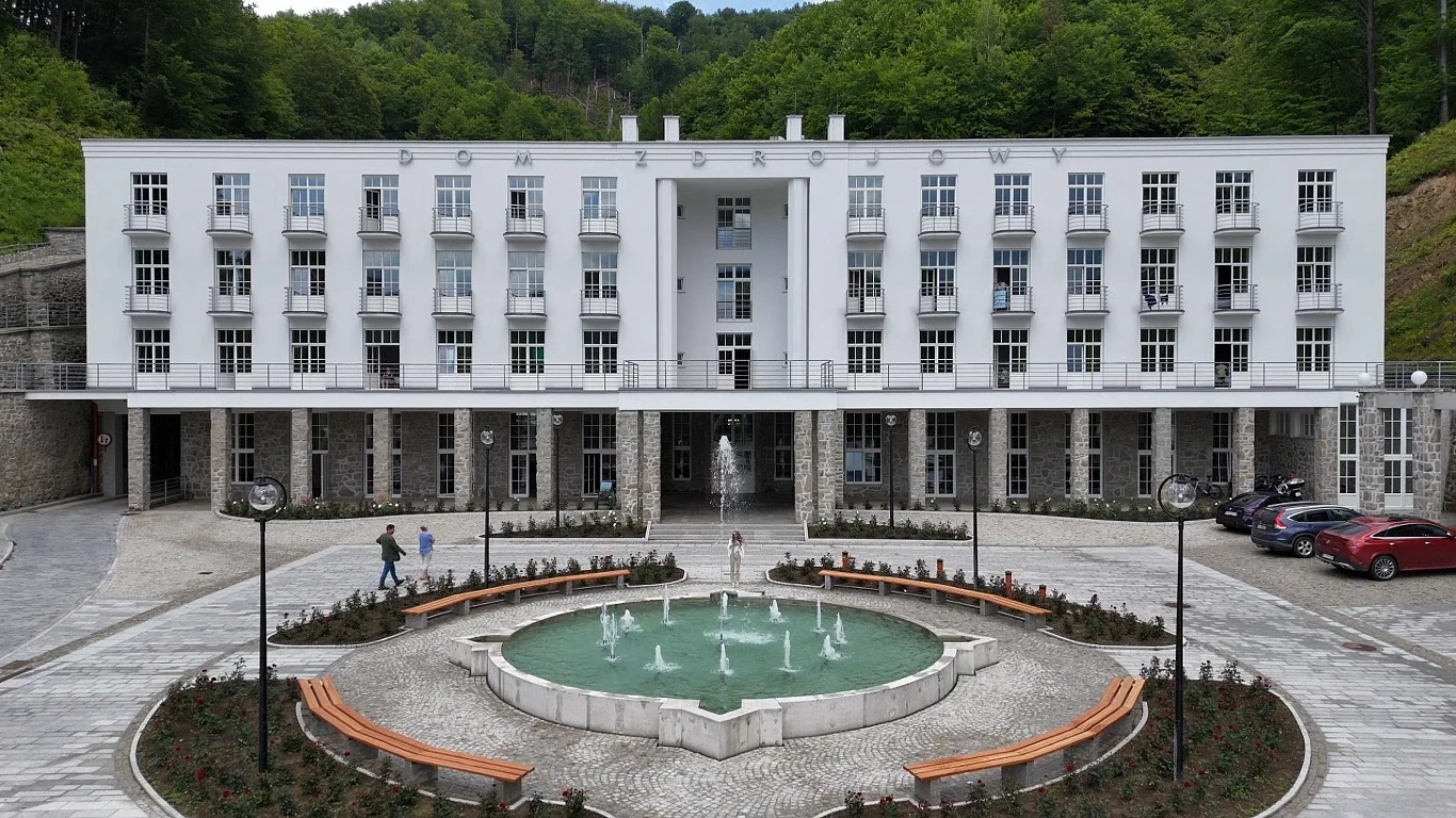Budynek o nowoczesnej, białej elewacji z licznymi balkonami, otoczony zielenią, na pierwszym planie okrągła fontanna z wodotryskami oraz ławkami ustawionymi półkoliście dookoła.
