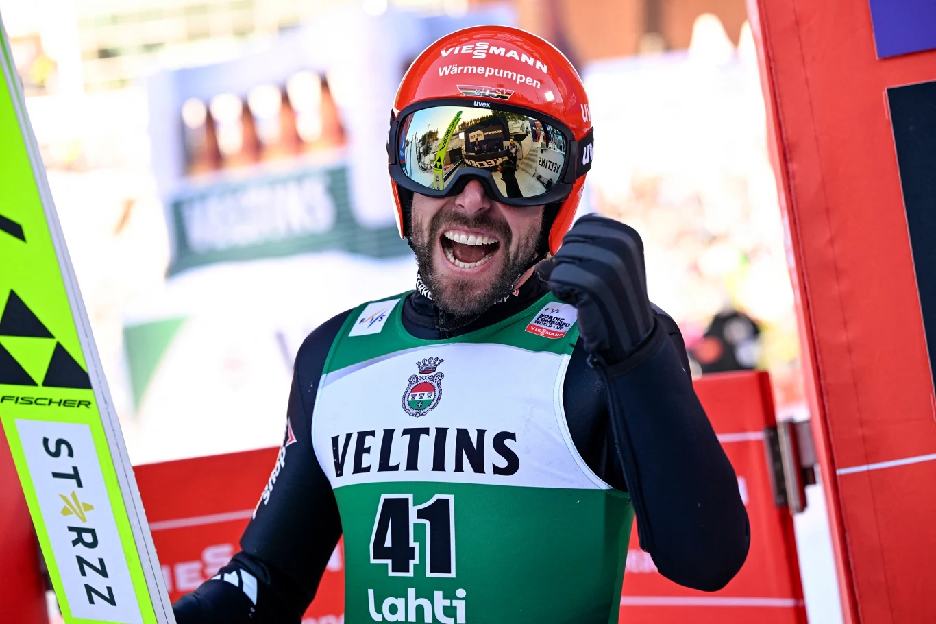 Skoczek narciarski w zielonym kombinezonie z numerem 41, kasku i goglach, trzymający nartę, wyraża radość po udanym skoku na zawodach, unosi zaciśniętą pięść w geście triumfu.