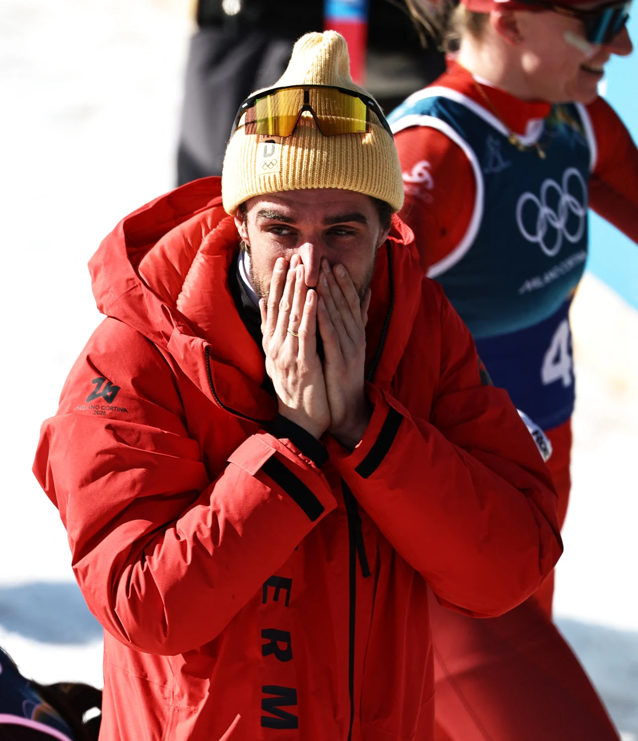 Mężczyzna w czerwonej kurtce i żółtej czapce z okularami na niej zakrywa dłonią usta w geście silnych emocji. W tle zawodniczka w niebiesko-czerwonym stroju sportowym z widocznym logo olimpijskim.
