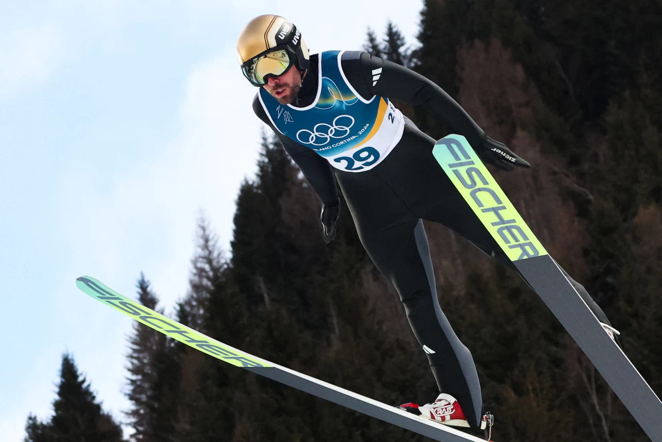 Skoczek narciarski w locie podczas zawodów olimpijskich, ubrany w czarny kombinezon, złoty kask i gogle, na nartach marki Fischer, w tle zarys ciemnych drzew.