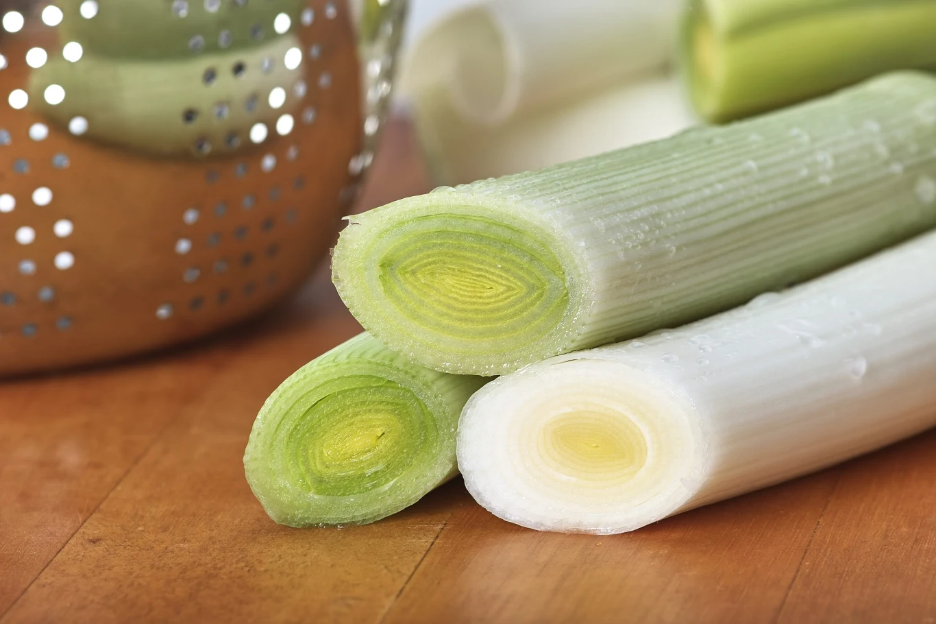 Świeżo pokrojone pory o jasnozielonym i białym wnętrzu leżą na drewnianym blacie, w tle widać metalowe sitko.