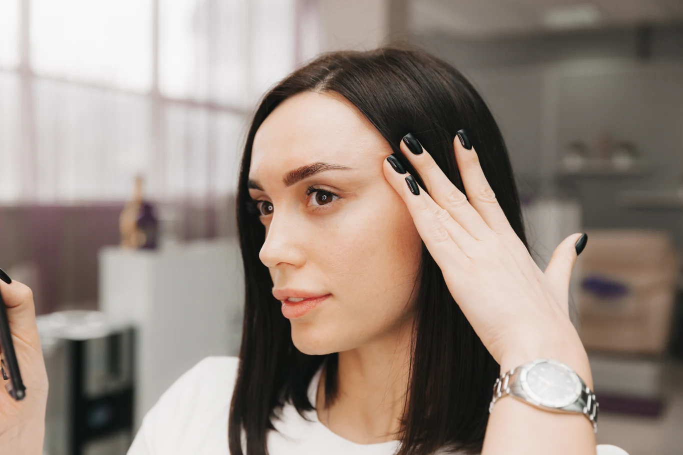 Młoda kobieta z ciemnymi, prostymi włosami trzyma dłoń przy twarzy, eksponując starannie pomalowane na czarno paznokcie; na nadgarstku widoczny srebrny zegarek, w tle jasne, rozmyte wnętrze.